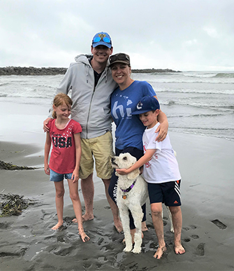 A man and woman are standing barefoot on a wet, sandy beach flanked by a young boy and girl on either side on a cloudy day. Small waves can be seen along the shore behind them and the boy is stroking the underside of a cream-colored dog’s chin.